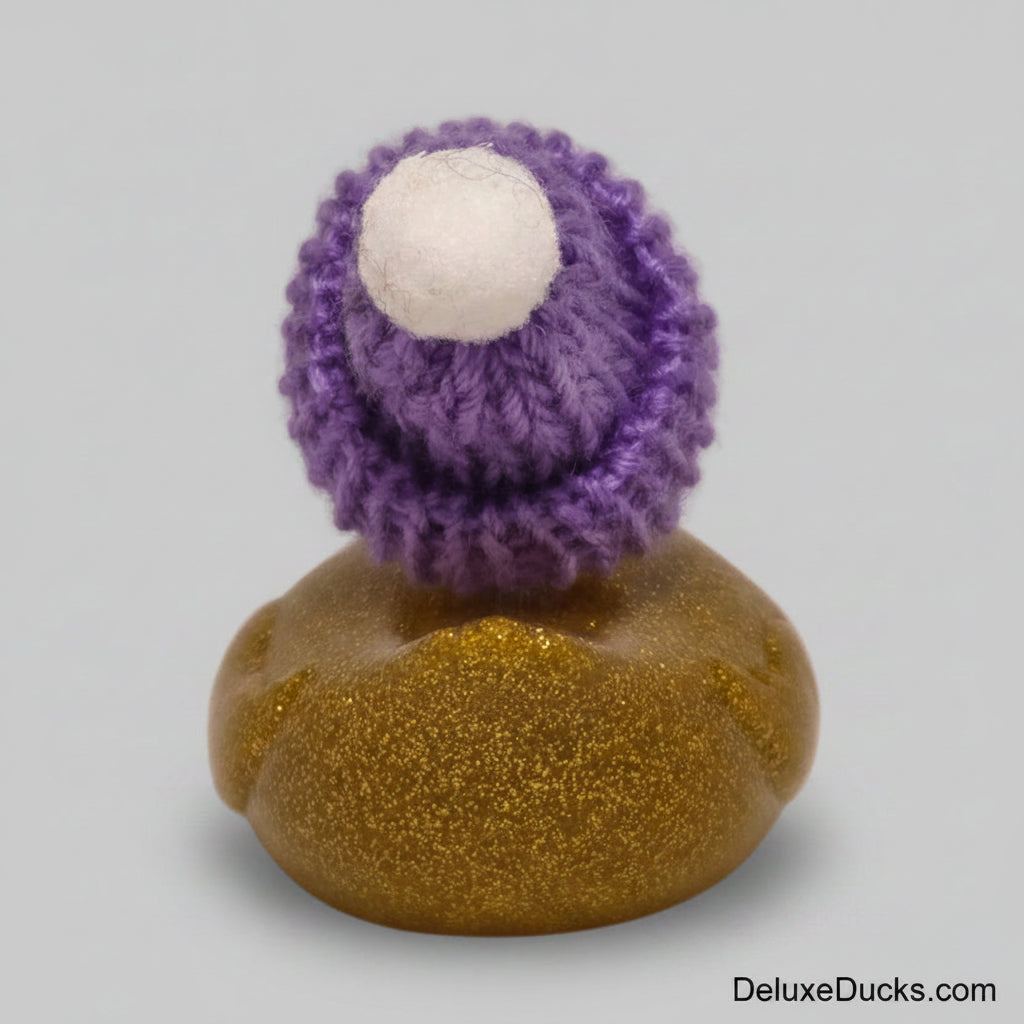 A small handmade decorative figure with a purple knitted hat, a white pom-pom, and a glittery gold body, photographed against a plain light background.
