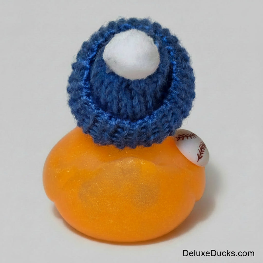 A small novelty baseball figurine featuring an orange glittery body, a blue knit beanie with a white pom-pom, and a miniature baseball accessory on its shoulder, set against a plain light background.