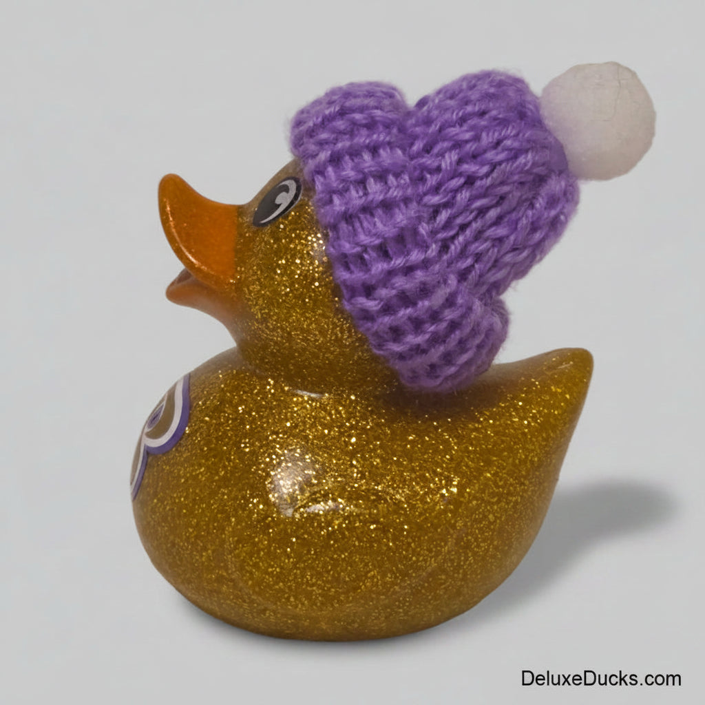 A glittery gold rubber duck wearing a purple knit hat with a white pom-pom, photographed against a plain light beige background.
