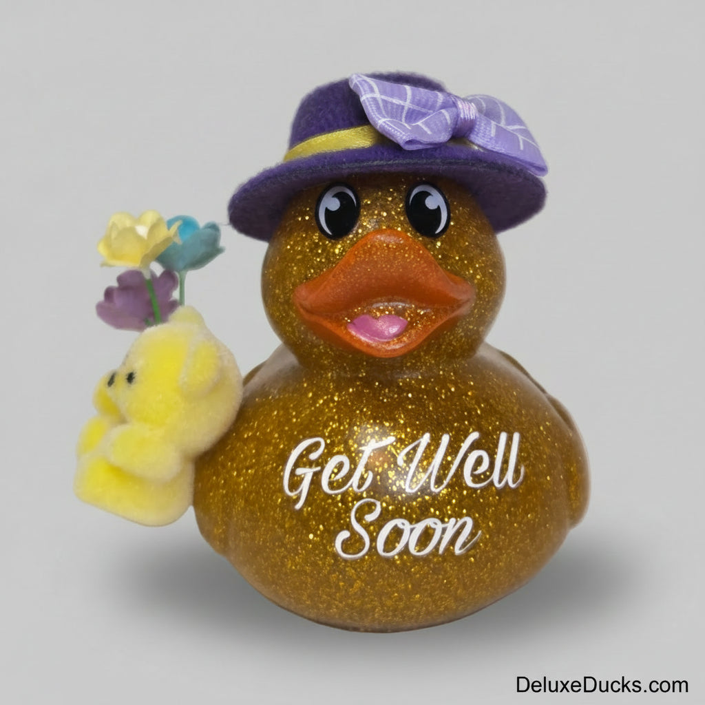 A glittery gold rubber duck figurine with 'Get Well Soon' written on its side, wearing a purple hat with a purple bow, and holding a small yellow plush chick with colorful flowers, set against a plain light background.