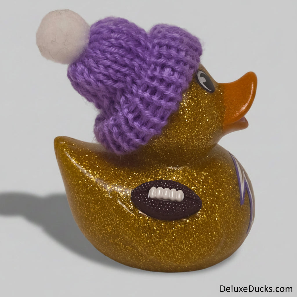 A glittery gold rubber duck wearing a purple knit beanie with a white pom-pom, featuring an orange beak, a purple football patch on its side, and a small blue and white logo.