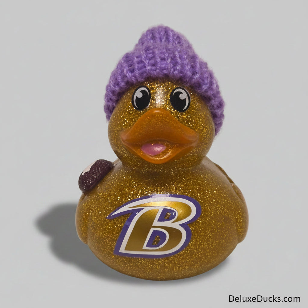 A glittery gold rubber duck with a purple knit beanie hat, featuring a large stylized letter 'B' logo on the front and a small purple pom-pom on top.